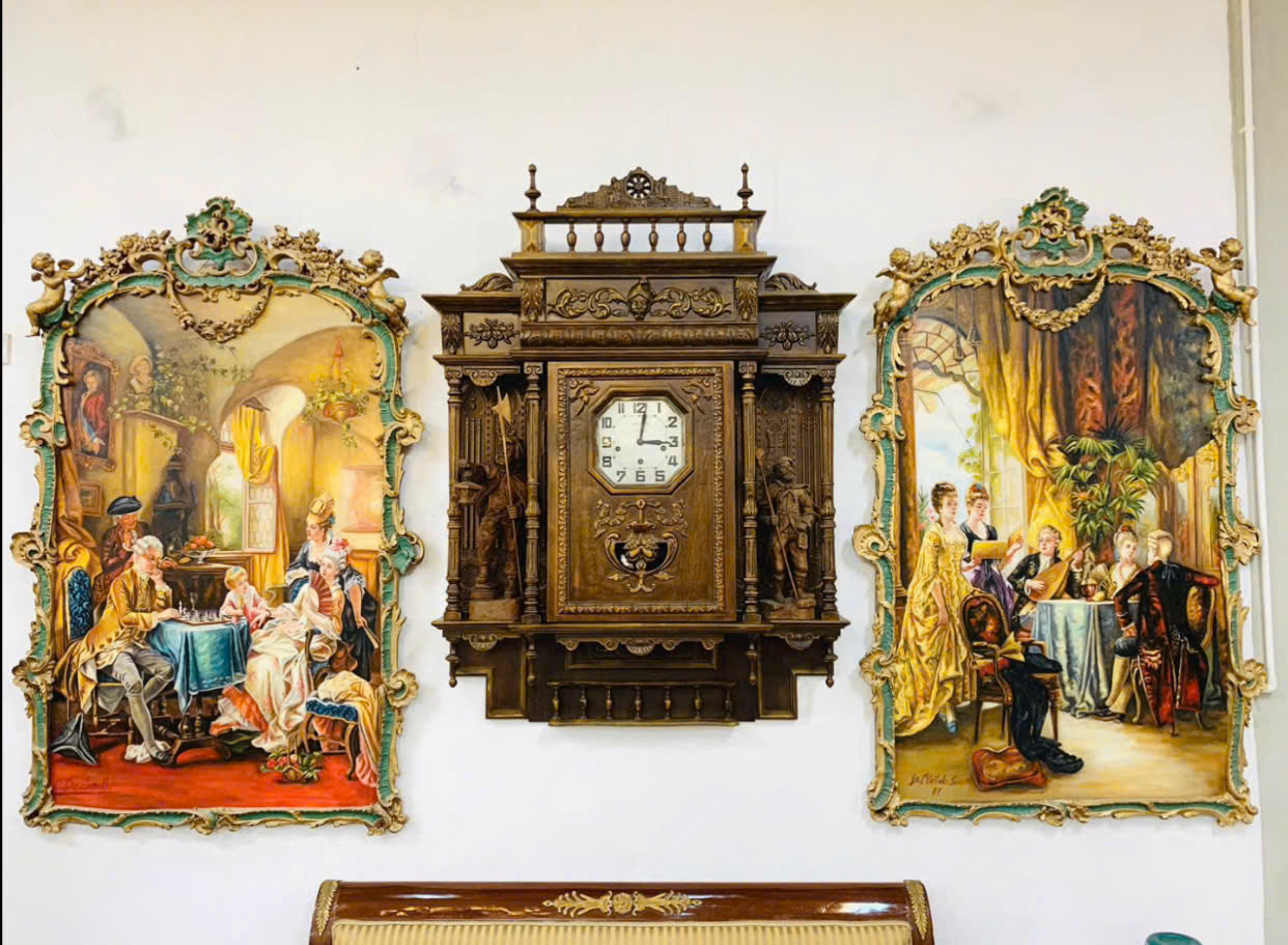 Đồng hồ treo tường gỗ cổ điển chạm khắc tinh xảo giữa hai bức tranh nghệ thuật phong cách châu Âu
